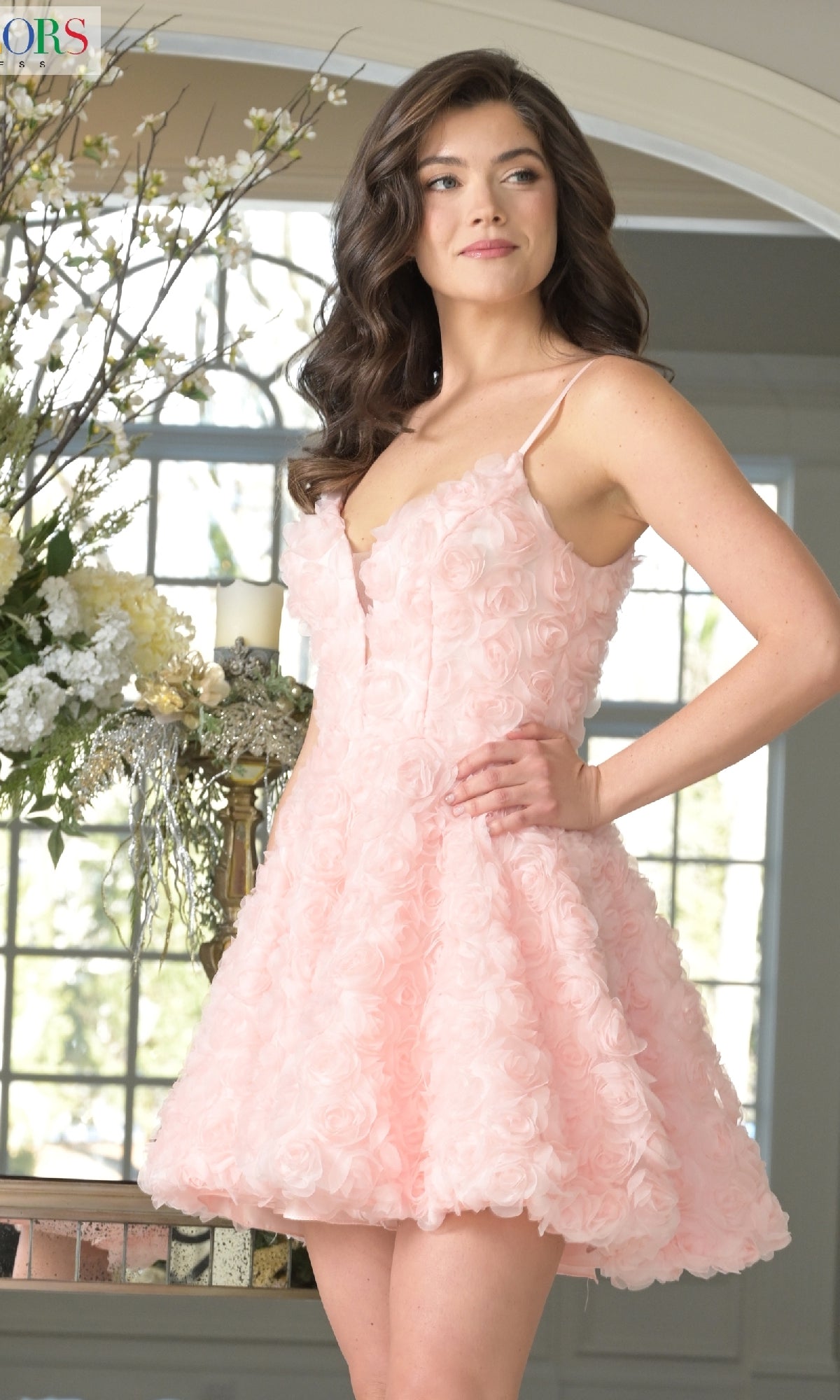 A woman poses indoors by a large window and floral display, wearing the Short Rosette A-Line Hoco Dress: Colors Dress 3672 in light pink, featuring sleeveless styling and delicate rosette details.